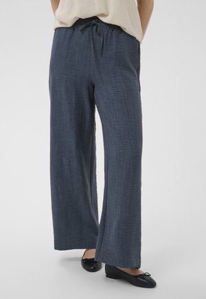 Femme portant un pantalon bleu marine texturé ample avec une taille à cordon de serrage et des ballerines noires, debout sur un fond blanc.