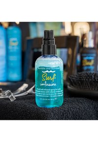 Spray Bumble and Bumble Surf Infusion in una bottiglia trasparente blu con pompa nera, etichettato per onde morbide e luminosità, 100 ml.