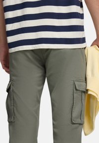 Pantalones cargo verde oliva con bolsillos laterales, combinados con una camiseta de rayas azul marino y blanca. Un suéter amarillo claro está colgado sobre un brazo.