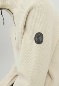 Crèmekleurige sweatshirt met ribbelstructuur en een ritssluiting bij de hals. Kenmerkt zich door een ronde zwarte patch met een wereldbolontwerp op de linkermouw.