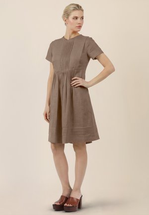 Donna in piedi con la mano sul fianco, che indossa un vestito marrone al ginocchio con dettagli plissettati e sandali marroni con zeppa e tacco.