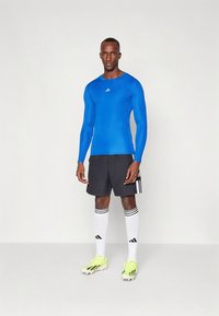 Kék hosszú ujjú sportfelső, fekete rövidnadrág, fehér zokni fekete csíkokkal, és élénksárga focicipő. Testhezálló kialakítás logó díszítésekkel.