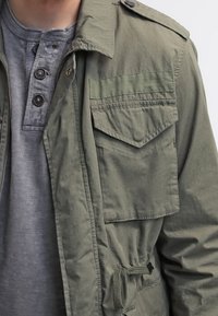 Mann trägt ein graues Henley-Shirt unter einer olivgrünen Jacke im Militärstil mit einem geknöpften Klappentaschen und elastischem Bund.