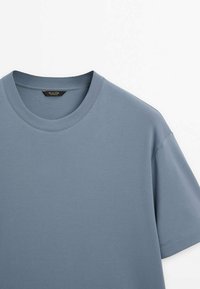 T-shirt uni à manches courtes avec col rond en bleu discret, présenté sur fond blanc, montrant l'encolure et la zone des épaules.