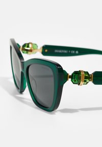 Ochelari de soare mari, de culoare verde, cu lentile întunecate. Au accente aurii și bijuterii din cristale verzi pe tâmple. Finisaj neted și lucios.