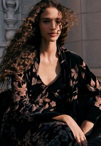 Juodas aksominis chalatas su rožinių gėlių raštais ir laisvai krentančiu dizainu, dėvimas ant juodos suknelės su gilia iškirpte.