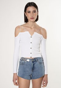 Top bianco a maniche lunghe con spalle scoperte, dotato di spalline argentate e chiusura frontale a bottoni; abbinato a pantaloncini in denim blu.