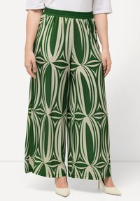 Grüne und beige weite Hosen mit einem auffälligen abstrakten Muster, elastischem Bund und leichtem Stoff. Kombiniert mit spitzen Schuhen.