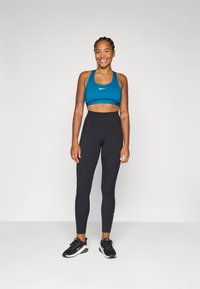 Donna sorridente in un reggiseno sportivo turchese e leggings neri, in piedi con sicurezza con le mani ai fianchi, su uno sfondo neutro.