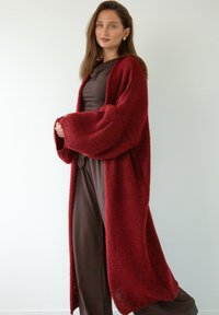 Burgundowy dziergany kardigan z oversizedowymi rękawami, teksturowany materiał, otwarty przód i długi fason, noszony na brązowy strój.