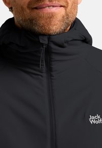 Veste légère noire avec une fermeture éclair avant, capuche ajustable et texture douce. Présente un petit logo blanc sur la poitrine.