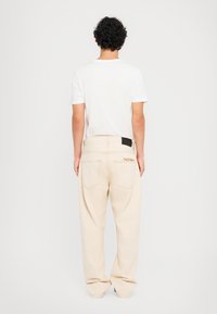 Pantaloni beige dal taglio rilassato, con due tasche posteriori e dettagli di cucitura discreti. Etichetta nera in vita. Maglietta bianca semplice.