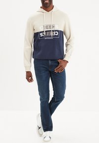 Sudadera con capucha en beige y azul marino con un bolsillo frontal, que presenta un texto destacado "CREEKS". Combinada con jeans oscuros y zapatillas blancas.