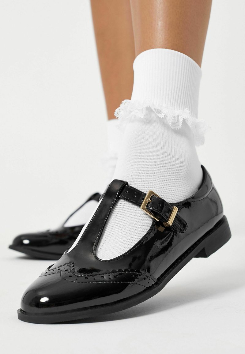 Svarta patentläderskor med T-bar-design, dekorativa broguedetaljer, ett guldspänne och tillsammans med en vit volangstrumpa.