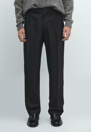 Pantaloni neri con un design frontale plissettato, caratterizzati da una vestibilità sartoriale e una texture liscia. Indossati con scarpe nere lucide, mostrati dalla vita in giù.