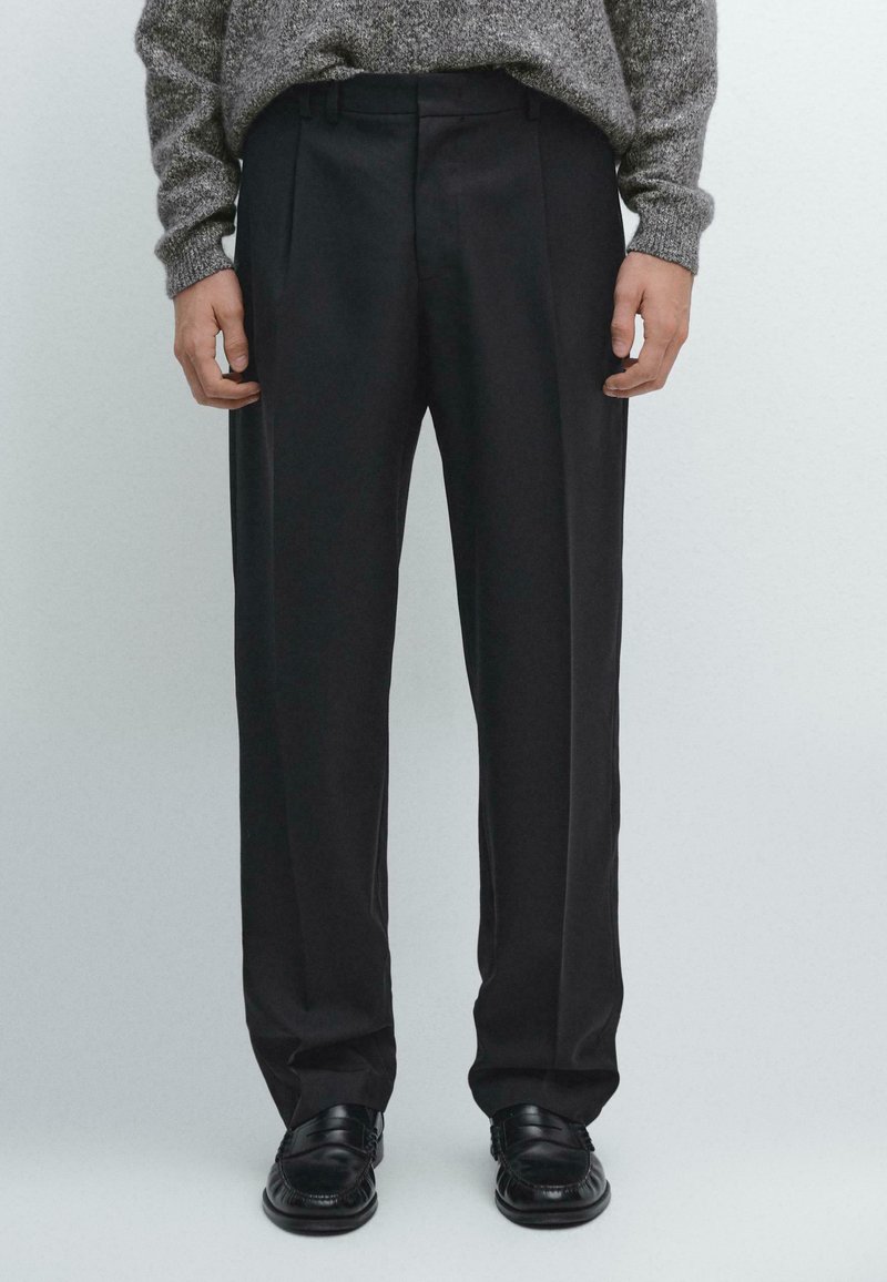 Pantalons noirs avec un design à plis sur le devant, présentant une coupe ajustée et une texture lisse. Portés avec des chaussures noires brillantes, présentés de la taille vers le bas.