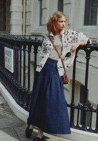 Balta megzta kardiganas su raštuotais akcentais, bežinis marškinėlis, ilga mėlyna sijonas, juodi loafersai, stovinti prie juodos kalimo geležies turėklų.