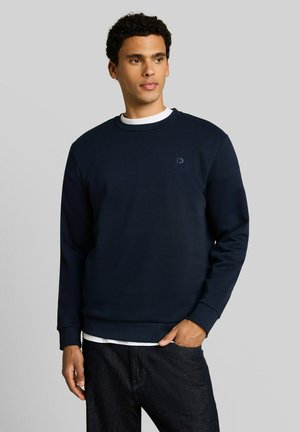 Man die een navy blauwe sweatshirt draagt over een wit overhemd en zwarte jeans, staand met één hand in zijn zak tegen een grijze achtergrond.