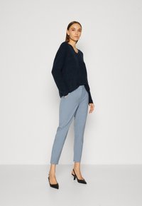 Marineblauwe gebreide trui met een brede halslijn, gecombineerd met lichtblauwe, op maat gemaakte broek met een slim fit en subtiele zakdetails. Zwarte hakken completeren de outfit.