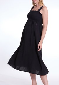 Schwarzes, ärmelloses Schwangerschafts-Kleid mit gerafftem Oberteil, fließendem Rock und verstellbaren Trägern. Leichter Stoff mit einer strukturierten Oberfläche.