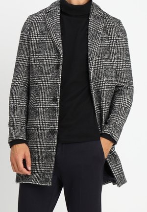 Homme portant un manteau à carreaux noir et blanc sur un col roulé noir avec une main dans la poche, sur un fond uni.