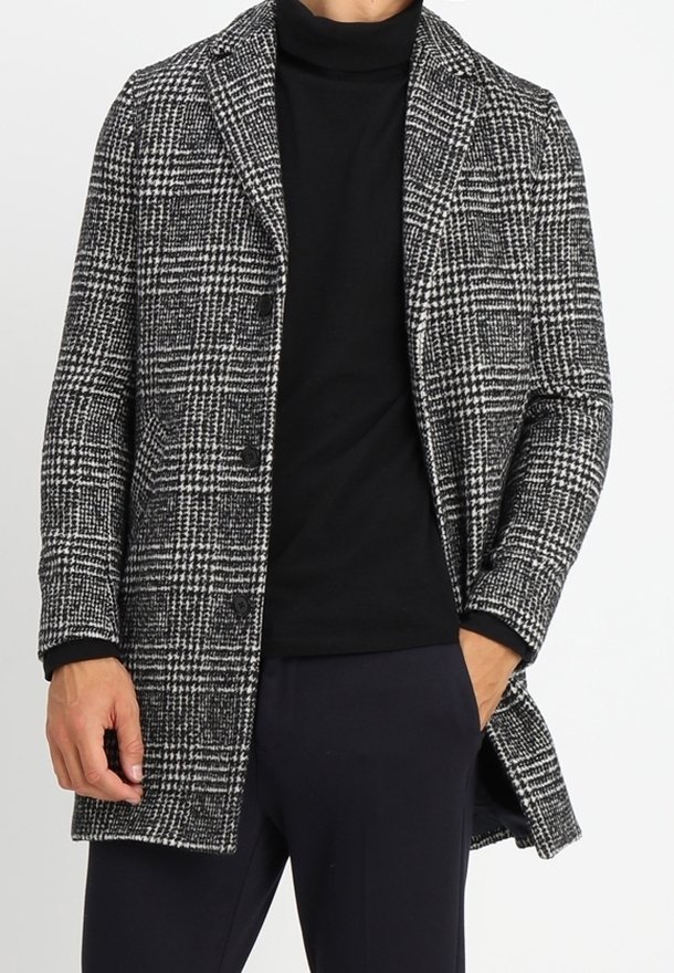 Homme portant un manteau à carreaux noir et blanc sur un col roulé noir avec une main dans la poche, sur un fond uni.