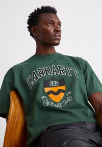 Zielona bawełniana koszulka z logo "CARHARTT", pomarańczową i czarną grafiką w kształcie tarczy oraz białym tekstem na wstążce. Krótkie rękawy i okrągły dekolt.