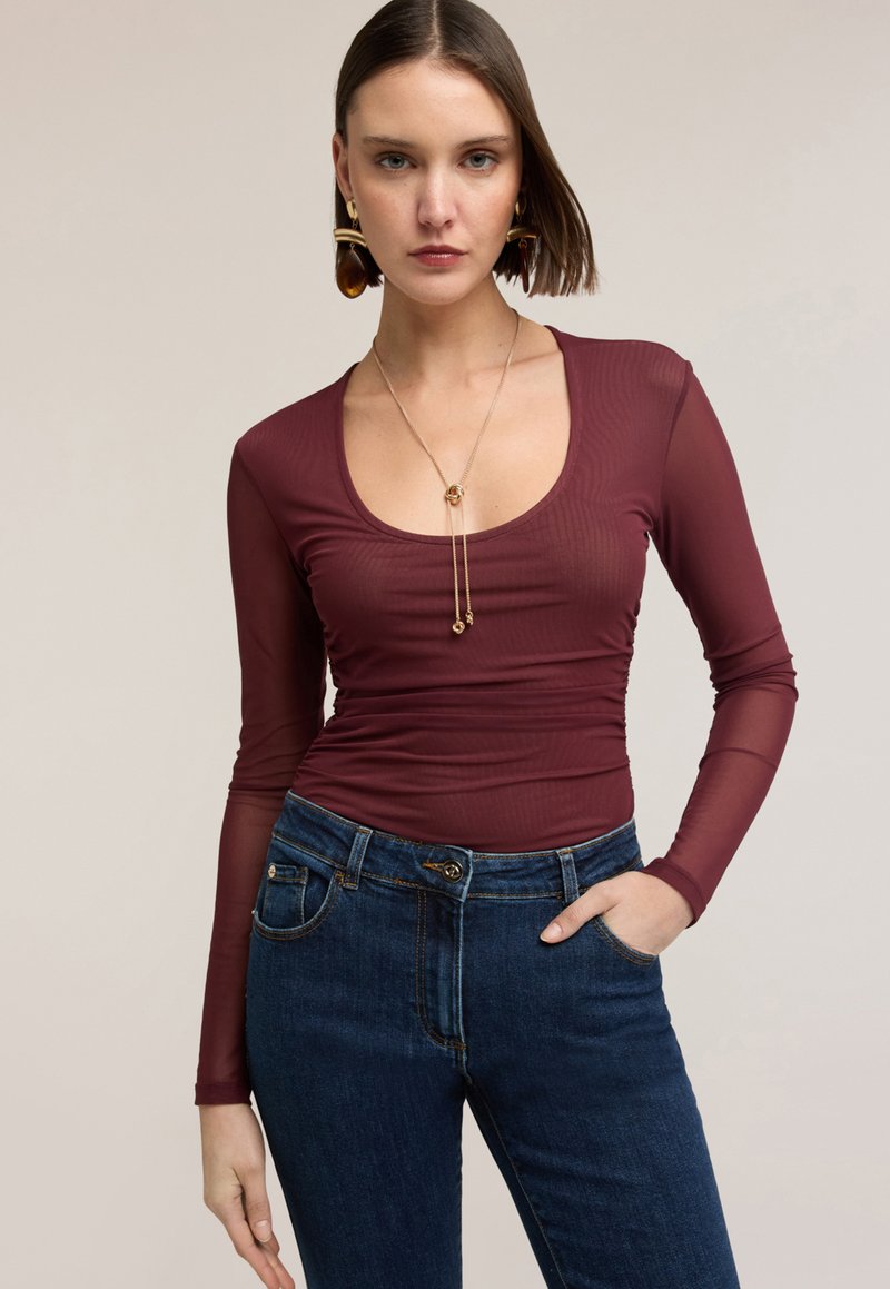Mujer vestida con una blusa ajustada de manga larga color burdeos y jeans azules de tiro alto, accesorios con un collar dorado y grandes pendientes.