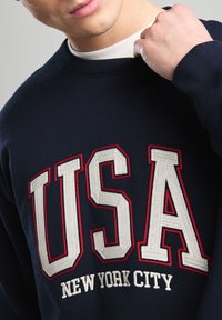 Navy sweatshirt featuring large, white and red embroidered "USA" and "NEW YORK CITY" text. Crew neck design with soft texture.