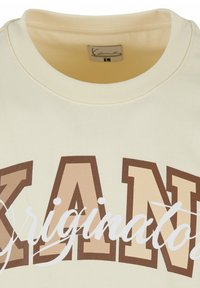 Cream-colored sweatshirt with a round neckline. Features large brown and beige graphic text "KANE" and smaller white cursive "Original." Soft fabric.