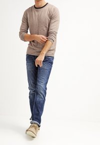 Hombre arremangándose las mangas de un suéter beige, vistiendo unos jeans azules y zapatos casuales color beige, de pie contra un fondo blanco.