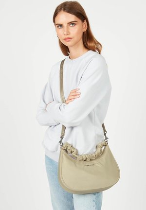 Borsa a spalla in pelle beige con bordo arricciato, forma curvata e tracolla staccabile con logo, tenuta da un modello in abbigliamento casual.