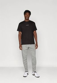 Μαύρο βαμβακερό μπλουζάκι με μικρό λευκό λογότυπο στο στήθος, συνδυασμένο με ανοιχτό γκρι φόρμα και λευκά αθλητικά παπούτσια, σε ουδέτερο φόντο.