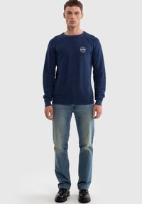 Tmavě modrý pletený svetr s žebrovanými manžetami a lemem, s kulatým výstřihem a malým bílým logem na prsou, doplněný světle modrými džíny.