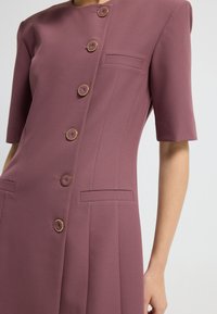 Un vestido a medida de tela malva presenta mangas cortas, seis botones circulares y dos bolsillos frontales con un diseño limpio y estructurado.