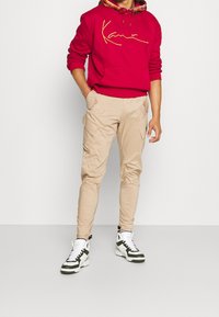 Sudadera roja con un logo en script amarillo, pantalones cargo beige con bolsillos laterales y zapatillas altas en blanco y negro con acentos verdes.