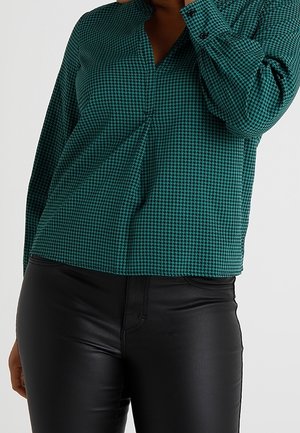 Kobieta w zielonej bluzce w kratę typu szkocka krata z długimi rękawami i czarnych skórzanych spodniach, pozująca z jedną ręką przy twarzy.