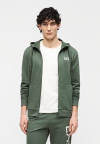 Donkergroene hoodie met rits, voorzak, capuchon met trekkoord en wit EA7-logo op de borst. Gecombineerd met bijpassende broek.