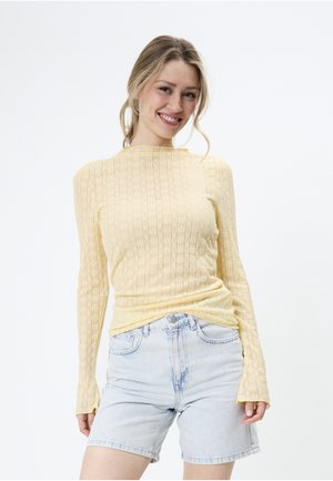 Frau lächelt, trägt ein hellgelbes, langärmeliges, strukturiertes Oberteil und hellblaue Jeans-Shorts vor weißem Hintergrund.