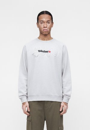 Muž s vitiligom, ktorý nosí svetlosivú mikinu Quiksilver a olivové cargo nohavice, stojaci pred jednoliatym bielym pozadím.