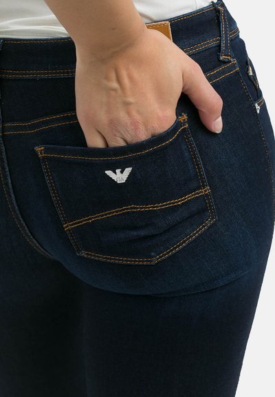 Tumedat sinised denim-püksid kontrastsete oranžide õmblustega. Omab väikest taskut koos hõbedase logoga, näidates kätt, mis on sisestatud tagataskusse.