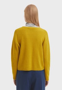 Maglione giallo senape con una finitura testurizzata, spalle scese e un taglio corto, abbinato a un dolcevita grigio sotto.