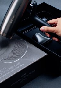 Main tenant la poignée noire d'un sèche-cheveux, partiellement soulevée d'une boîte noire contenant deux embouts et un emballage portant la mention « ghd speed ».