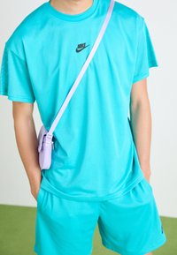 Osoba oblečená v tyrkysovém síťovaném sportovním outfitu Nike s odpovídajícími šortkami a levandulkovou krosbody taškou před jednobarevným pozadím.