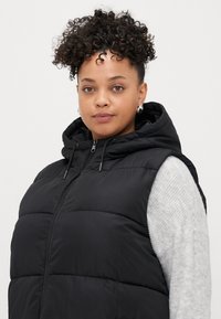 Melnā iešūšanas vesta ar kapuci, rāvējslēdzēju un priekšējām kabatām. Uzvilkts pāri gaiši pelēkam džemperim. Iezīmē gludu tekstūru un regulējamu kapuci.