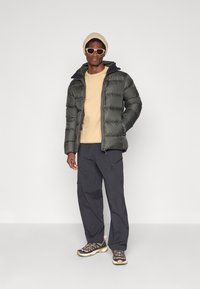 Homem com um gorro bege, óculos de sol brancos, sweatshirt bege, casaco acolchoado verde escuro, calças cargo pretas e sapatos de montanha multicoloridos, em pé.