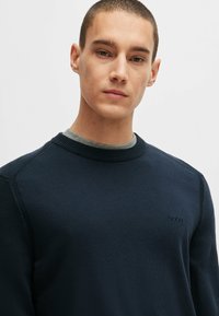 Marineblauwe gebreide sweatshirt met een ronde halslijn en geribde boorden. Bevat een tonale logo op de borst en een gelaagd grijs T-shirt eronder.