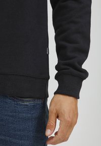 Felpa nera con polsini e orlo a coste, caratterizzata da un'etichetta sul lato. Il tessuto sembra morbido e testurizzato. Indossata con jeans blu.