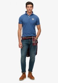 Polo bleu avec col, logo brodé, chemise en flanelle à carreaux nouée autour de la taille, jean bleu, bottes en cuir marron avec lacets.