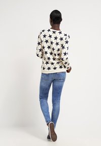Sudadera crema con patrón de estrellas marinas, cuello redondo y mangas largas, combinada con jeans ajustados de color azul claro y zapatos casuales.